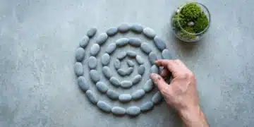 A human hand arranging river stones into a geometric spiral on concrete, next to a moss terrarium, symbolizing holistic sustainable living principles beyond simple recycling.